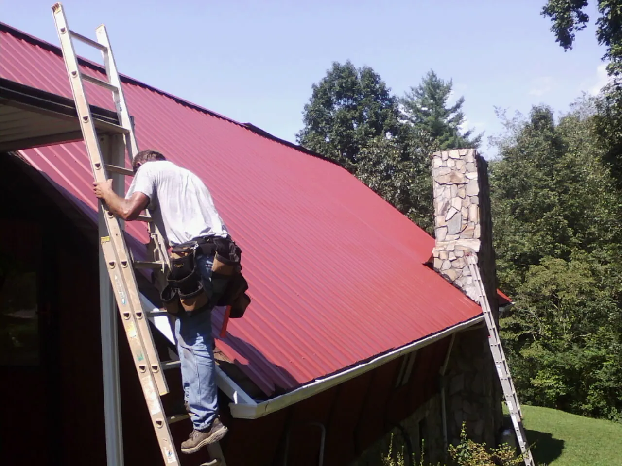Roofer on a ladder beside a red metal roof and stone chimney — Soffit & Fascia Repair by Roofing Brothers in Jay